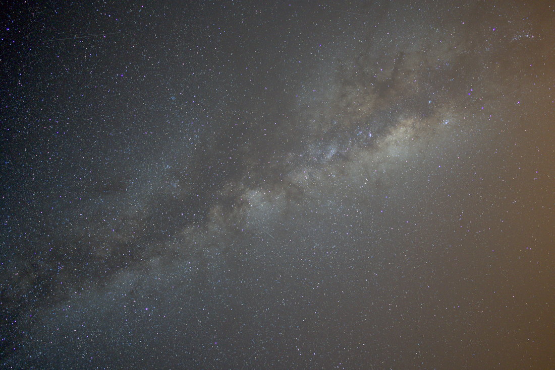 A single 20-second frame of the Milky Way at 24 mm and f/1.4: the galactic core crossing the frame diagonally, a faint satellite track at top left, and an orange glow of light pollution in the bottom-right corner