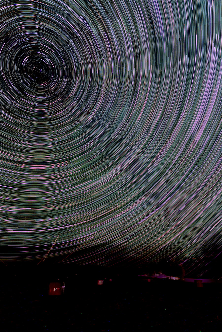 A star-trail stack: concentric arcs turning about the celestial pole over a dark tree line and hay bales in a field