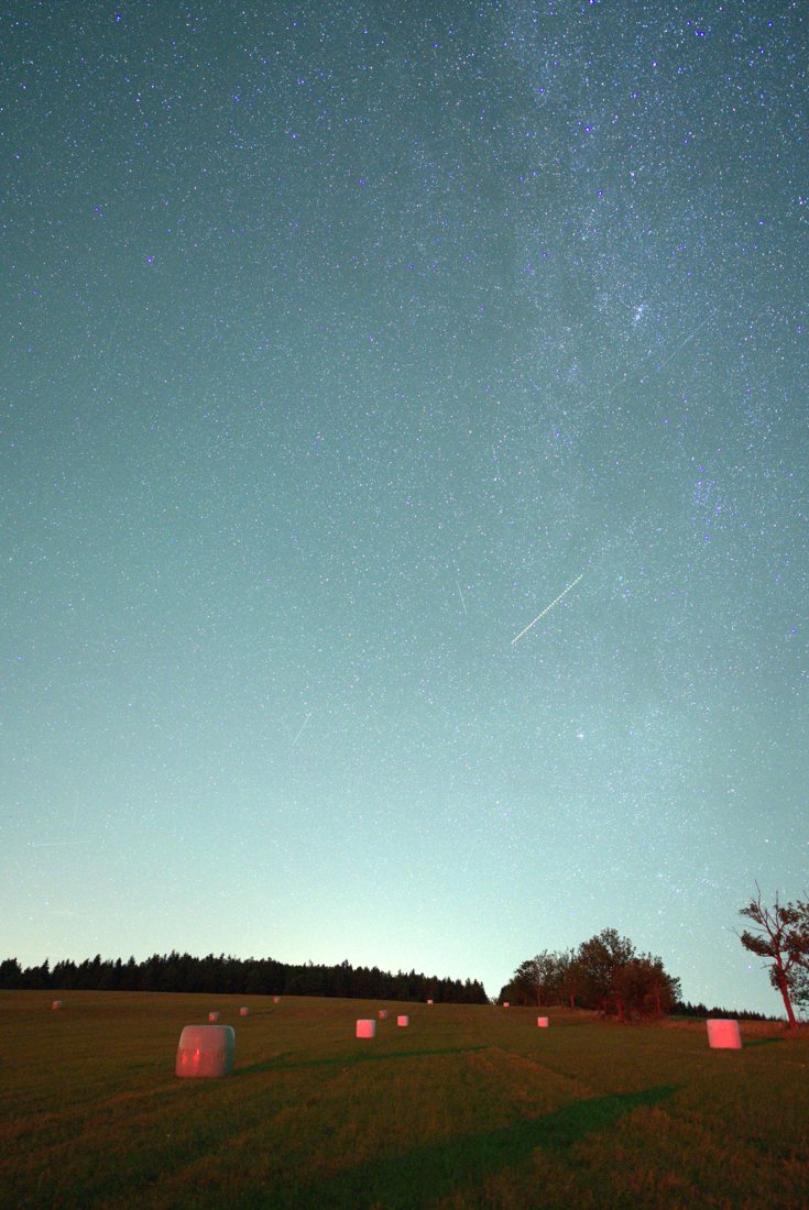A rural night sky glowing teal from light pollution, only a scatter of bright stars and the Milky Way barely visible, over a field of hay bales lit red